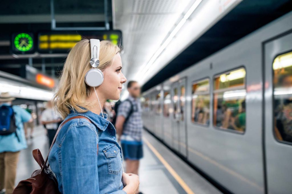 Beautiful young blond woman in denim shirt with earphones, standing at the underground platform, waiting to enter a train listening to Audio Lessons on Insurance from insuranceexampodcast.com.